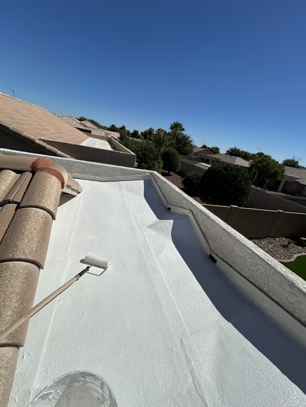 After: the same flat roof section freshly coated in white waterproof roof coating, with the paint roller on the roof